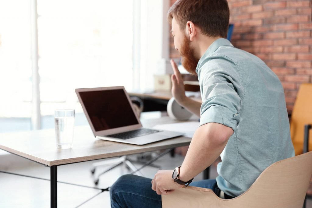 Image of a smiling young man sitting at a desk waving at a laptop. If you're struggling to go to in-person therapy, learn how telehealth in Denver, CO can help you!
