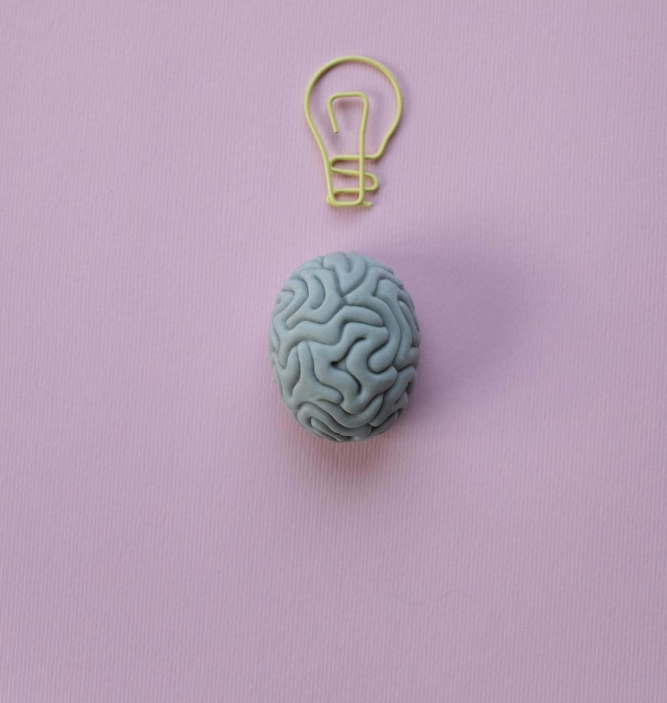 A small, sculpted brain made of blue clay sits on a pastel pink background, with a yellow wire bent into the shape of a lightbulb above it. This could represent the importance of brain health and overcoming trauma with a neurofeedback therapist in Englewood, CO. Search for trauma therapy in Greenwood Village, CO and how neurofeedback therapy can help.