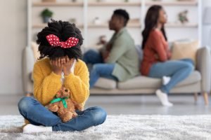 A young girl with curly hair, wearing a yellow sweater and a red polka-dot bow, sits on the floor holding a teddy bear while covering her face. In the background, her parents sit on a couch with their backs turned to each other. This could represent the effects of childhood trauma that a neurofeedback therapist in Englewood, CO can help address. Learn more about trauma therapy in Englewood, CO and how neurofeedback therapy in Englewood, CO can offer support today. 