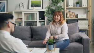 A woman with shoulder-length red hair and a striped shirt sits on a gray couch, smiling while talking to a man with a clipboard who is partially visible in the foreground. This could represent the support that neurofeedback therapist in Englewood, CO can offer. Search for trauma therapy in Greenwood Village, CO to contact a trauma therapist in Englewood, CO today. 