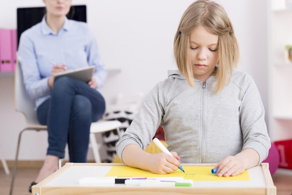 A girl draws with markers while a therapist observes in the background during autism testing in Greenwood Village, CO. Early evaluation for autism testing in Littleton, CO can help families better understand subtle signs related to autism in women Littleton, CO as they grow.