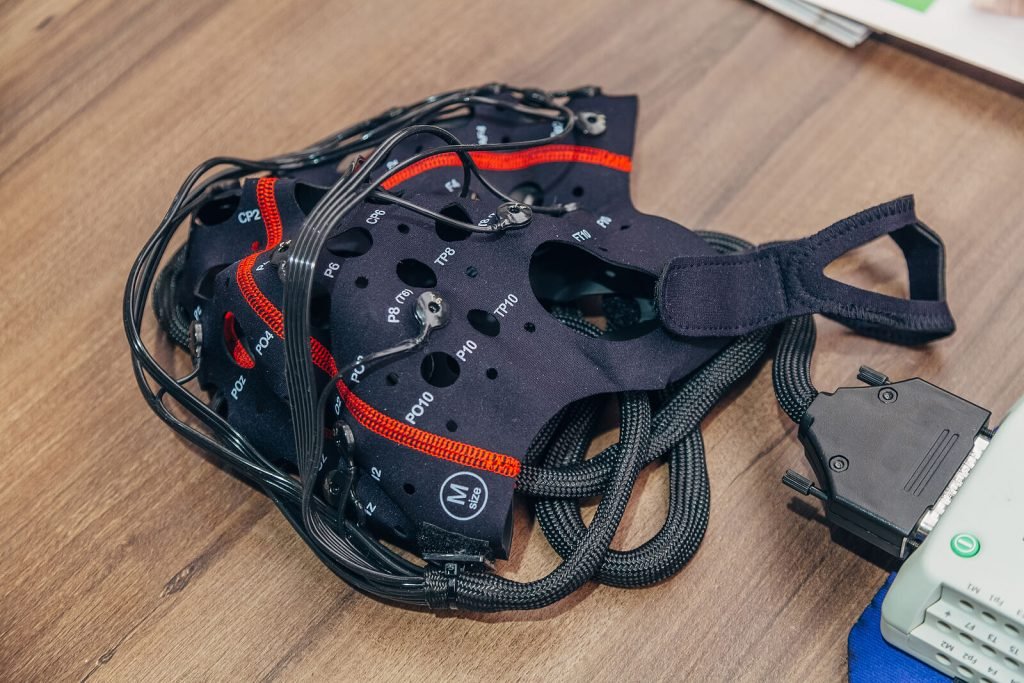 A close-up of a neurofeedback EEG cap laid flat on a wooden surface, showing the labeled sensor positions, wired electrodes, and connected amplifier device used to map and monitor brain activity. This is the same type of equipment used during a QEEG brain mapping appointment with a neurofeedback therapist in Englewood, CO — a painless, non-invasive first step toward understanding how your brain is functioning. For those searching for neurofeedback near me, this is what the process looks like up close before a single session even begins.